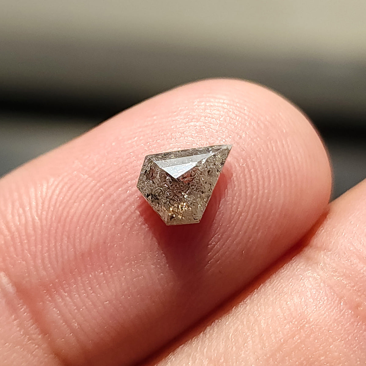 Rotated shield-shaped salt and pepper diamond on a fingertip, with a broad light reflection near the upper edge.