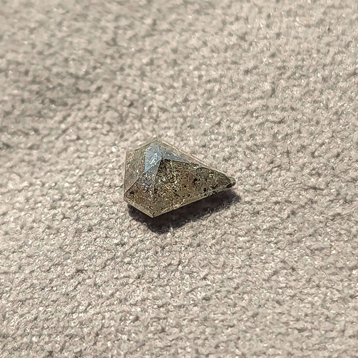 Angled shield-shaped salt and pepper diamond on a textured gray surface, with a shadow beneath the faceted crown.