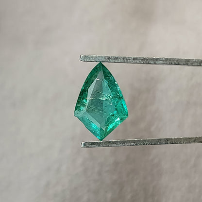 Face-up green shield-shaped emerald in tweezers against gray, with visible internal details and bright green edges.