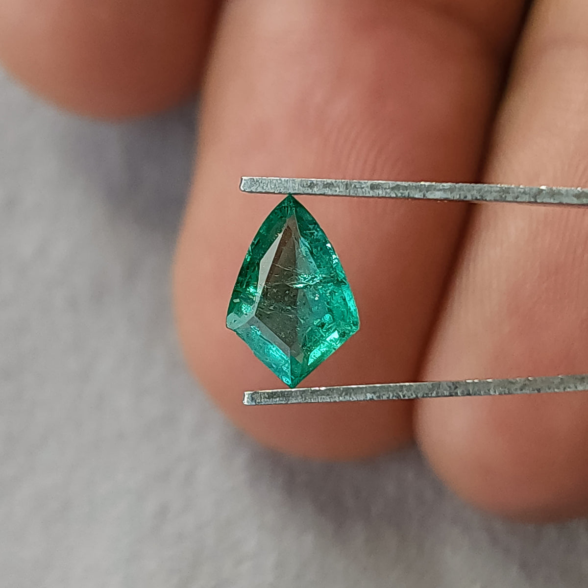 Green shield-shaped emerald in tweezers against fingers for scale, showing its front facets and dark internal details.