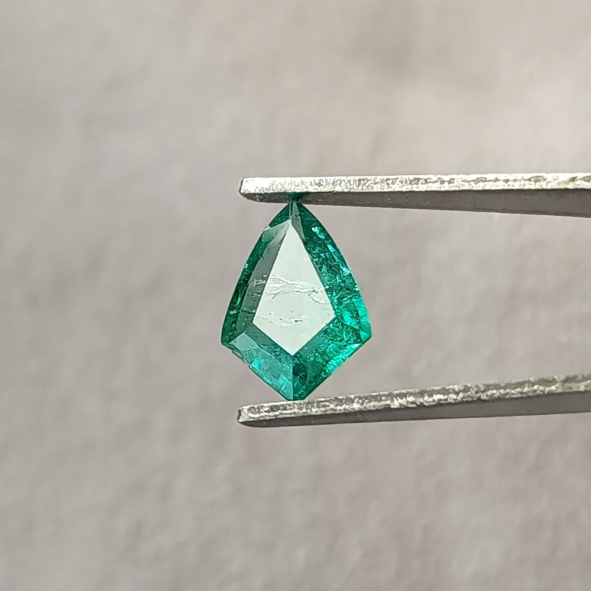 Table-side green shield-shaped emerald in tweezers, with a broad pale central reflection and green facet borders.