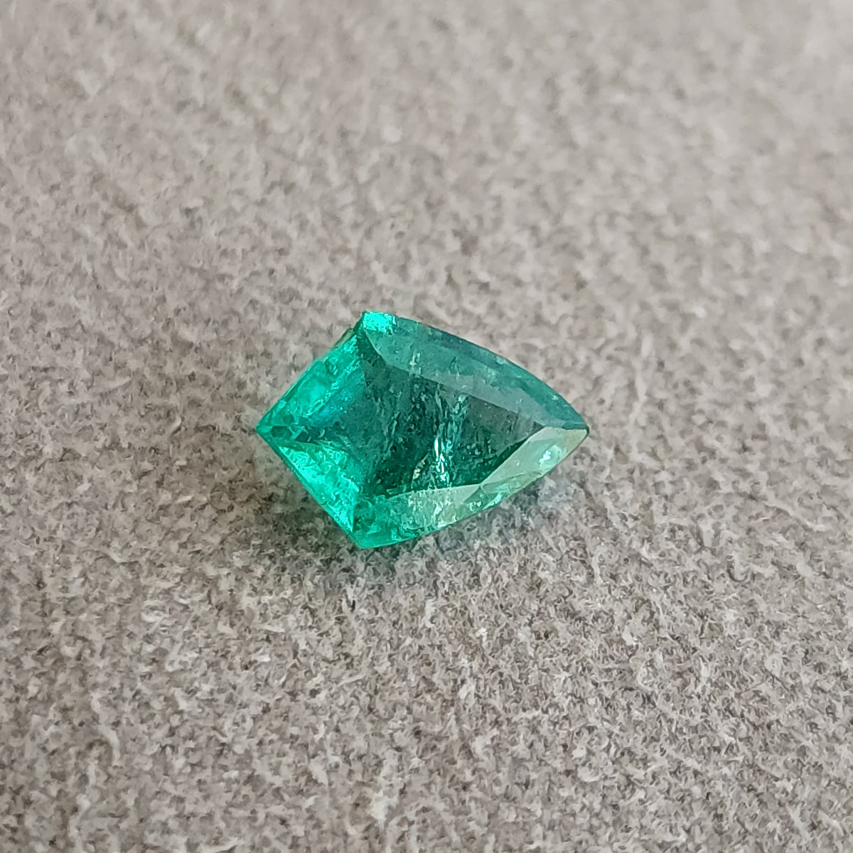 Angled table-side green shield-shaped emerald on a textured gray surface, with a dark center and bright green edges.