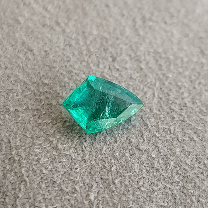 Angled table-side green shield-shaped emerald on a textured gray surface, with a dark center and bright green edges.