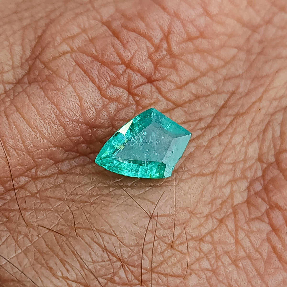 Green shield-shaped emerald resting on the back of a hand, with a pale table reflection and green border.