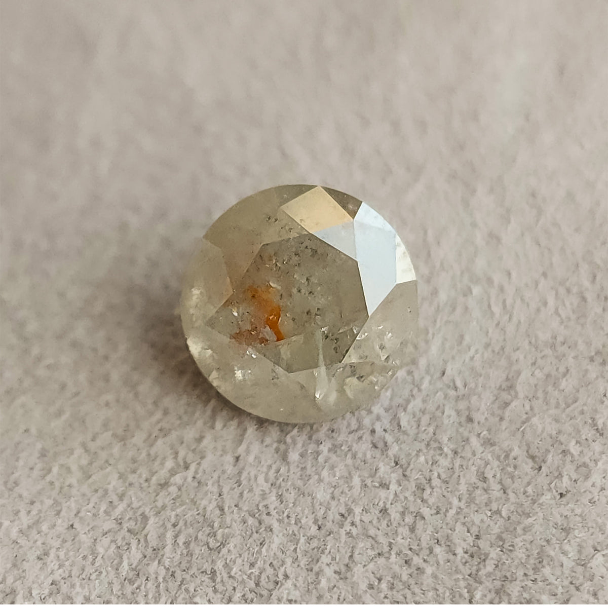 Round gray salt and pepper diamond resting face-up on a textured gray surface, with visible inclusions and a warm orange-brown internal feature.