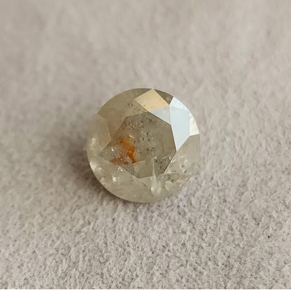 Round gray salt and pepper diamond resting face-up on a textured gray surface, with visible inclusions and a warm orange-brown internal feature.