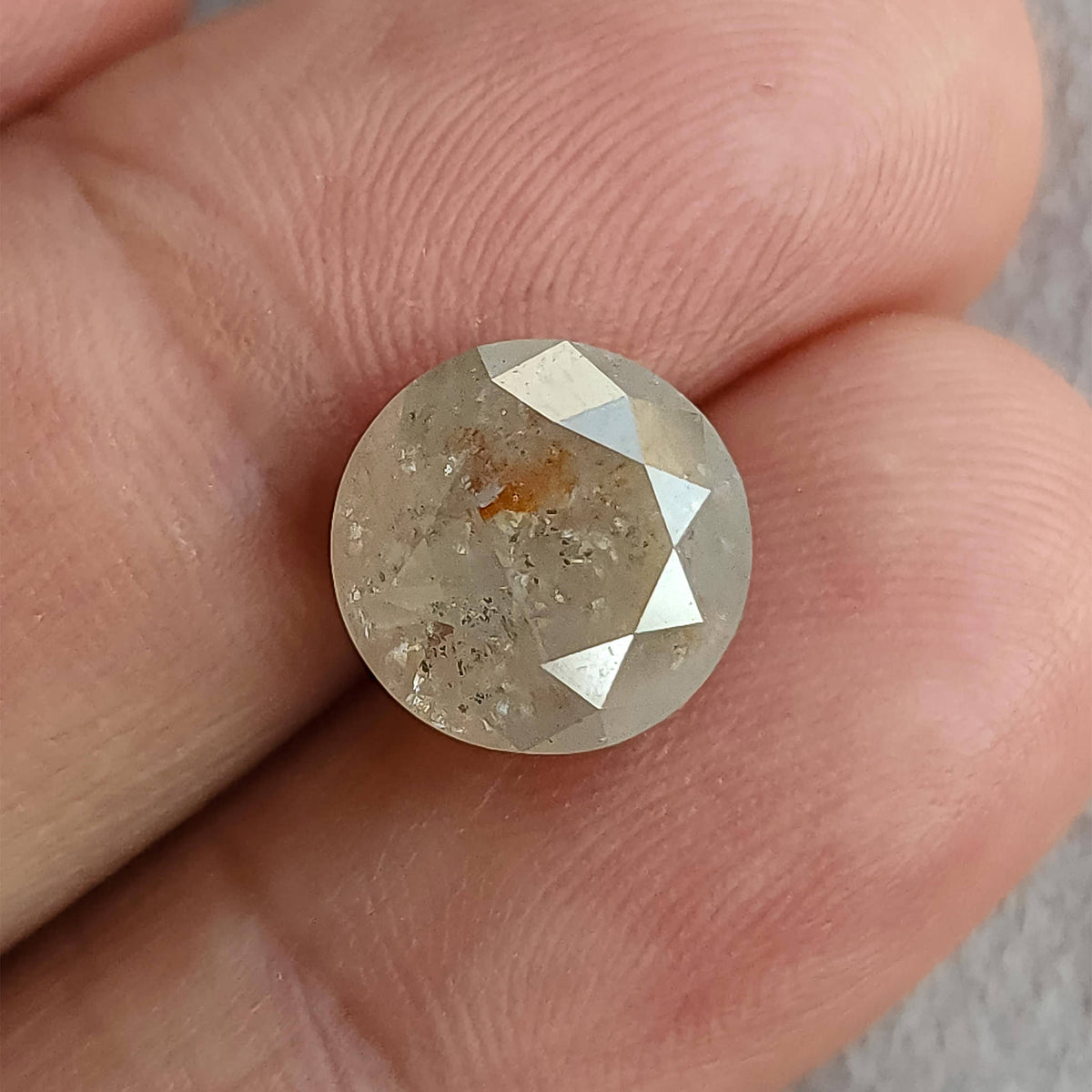 Round salt and pepper diamond against fingertips for scale, with front facets and a warm orange-brown inclusion visible.