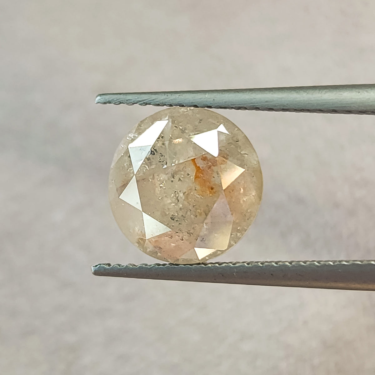 Round diamond held face-up in tweezers against gray, showing its brilliant-cut front facets, inclusions and a warm internal feature.