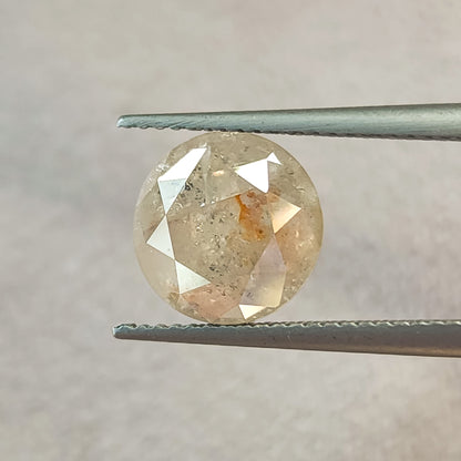 Round diamond held face-up in tweezers against gray, showing its brilliant-cut front facets, inclusions and a warm internal feature.