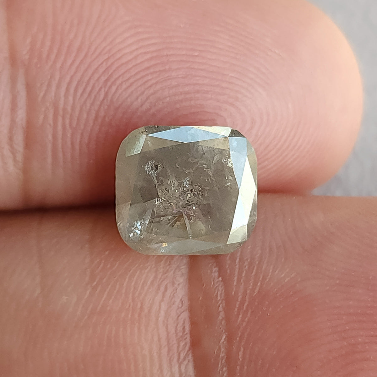Cushion-shaped diamond resting against fingertips for scale, with inclusions visible through the front table.