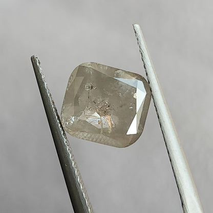 Cushion-shaped salt and pepper diamond held face-up in tweezers against gray, showing a broad table and visible inclusions.