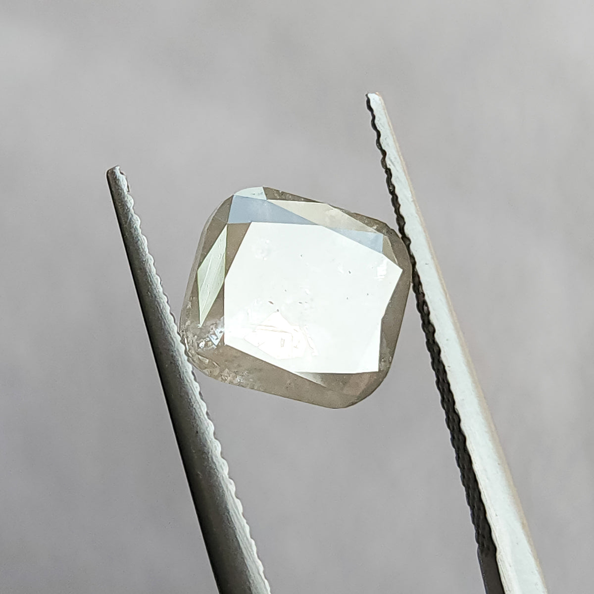The cushion diamond in tweezers at a changed angle, with a bright pale reflection across the front table and darker border facets.