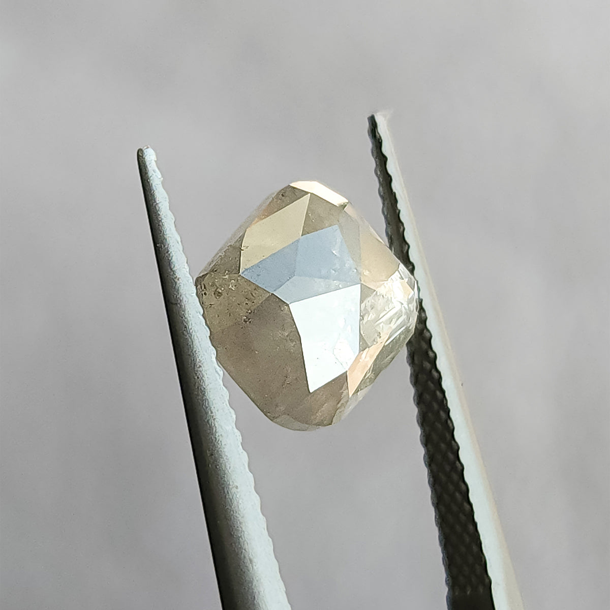 Opposing faceted side of the cushion diamond held in tweezers, with triangular pale reflections across its raised surface.