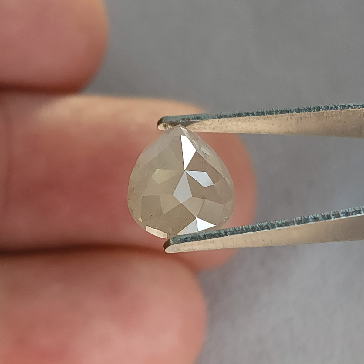 Rose-cut pavilion of a pear-shaped ice gray diamond in tweezers against fingers, showing converging triangular facets.