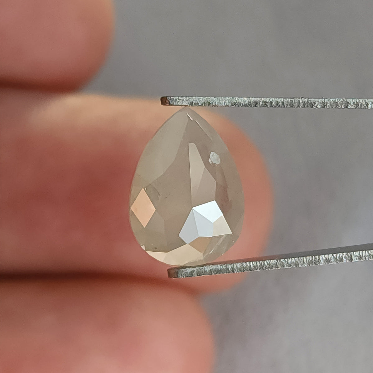 Pear-shaped ice gray diamond in tweezers, showing its rose-cut pavilion and a pale round feature near the upper right.