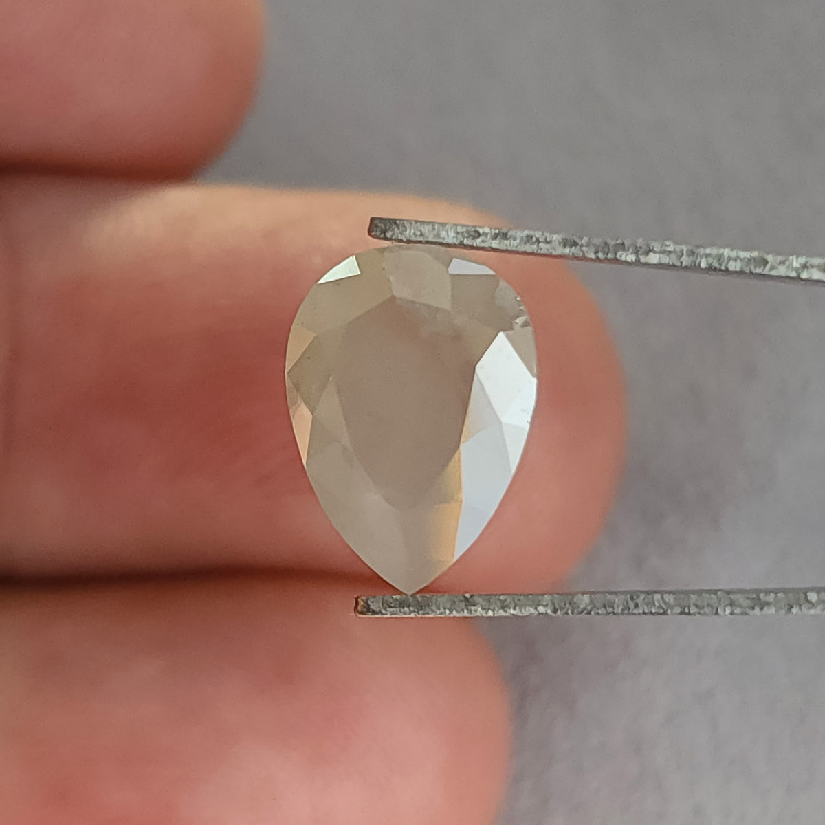 Table-side pear-shaped ice gray diamond in tweezers, with the point downward and a bright right-edge reflection.