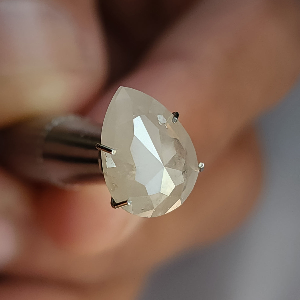 Angled rose-cut pavilion of a pear-shaped ice gray diamond in a temporary holder, with pale triangular reflections.