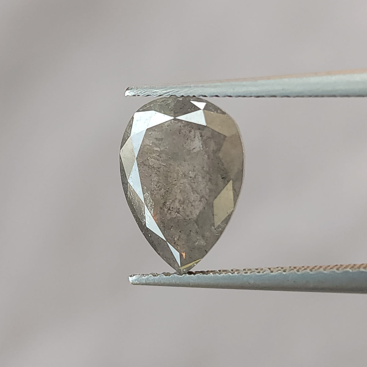Pear-shaped salt and pepper diamond held face-up in tweezers against gray, with its brilliant-cut table side and inclusions visible.