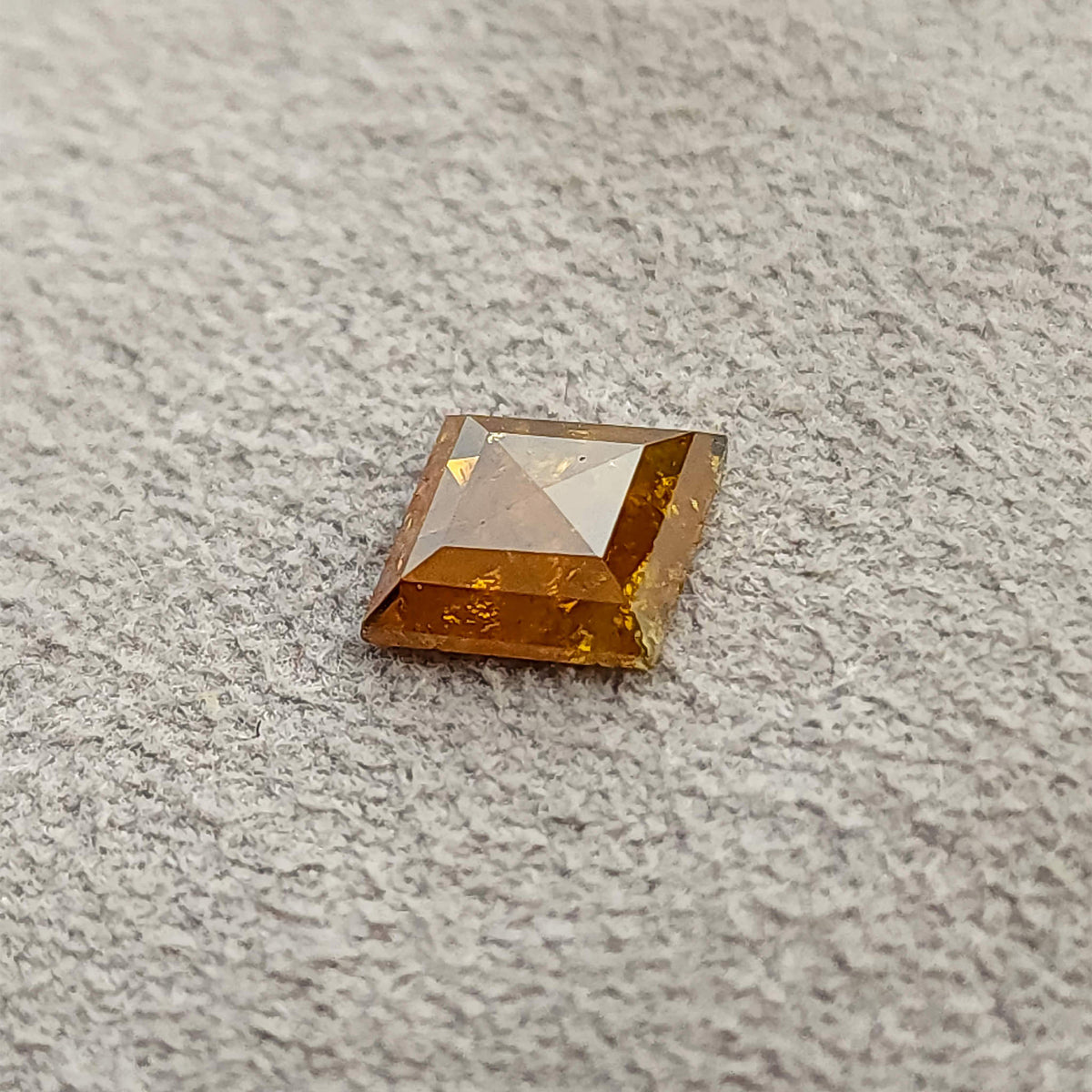 質感のあるグレーの表面に置かれたオレンジ色のカイト形ルースダイヤモンドを、低い位置から斜めに捉えた画像です。淡いファセットの反射と浅い形状が見えます。