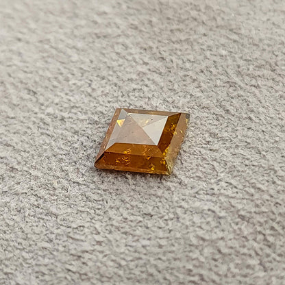 質感のあるグレーの表面に置かれたオレンジ色のカイト形ルースダイヤモンドを、低い位置から斜めに捉えた画像です。淡いファセットの反射と浅い形状が見えます。
