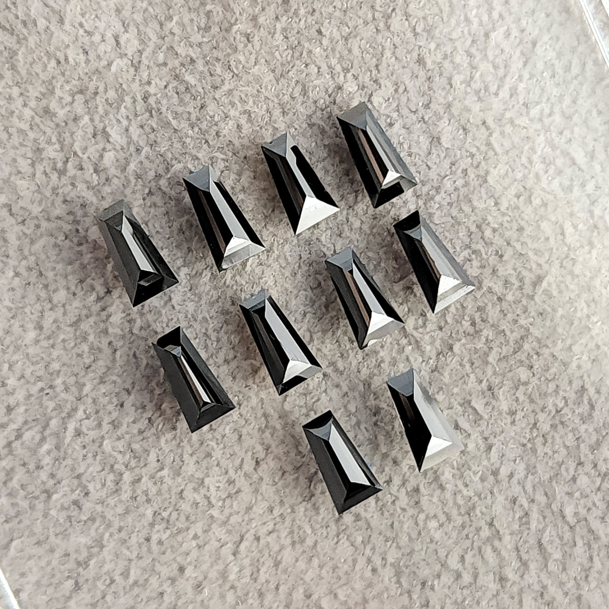 Black tapered baguette diamonds arranged on a pale textured surface, with bright narrow facet reflections.