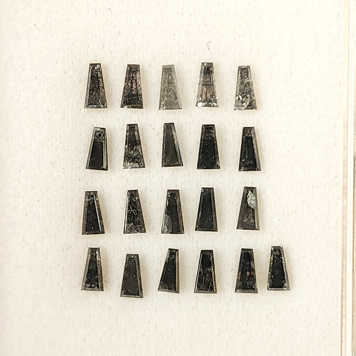 Group of natural salt and pepper tapered baguette diamonds arranged in a display box, showing mixed smoky gray tones.