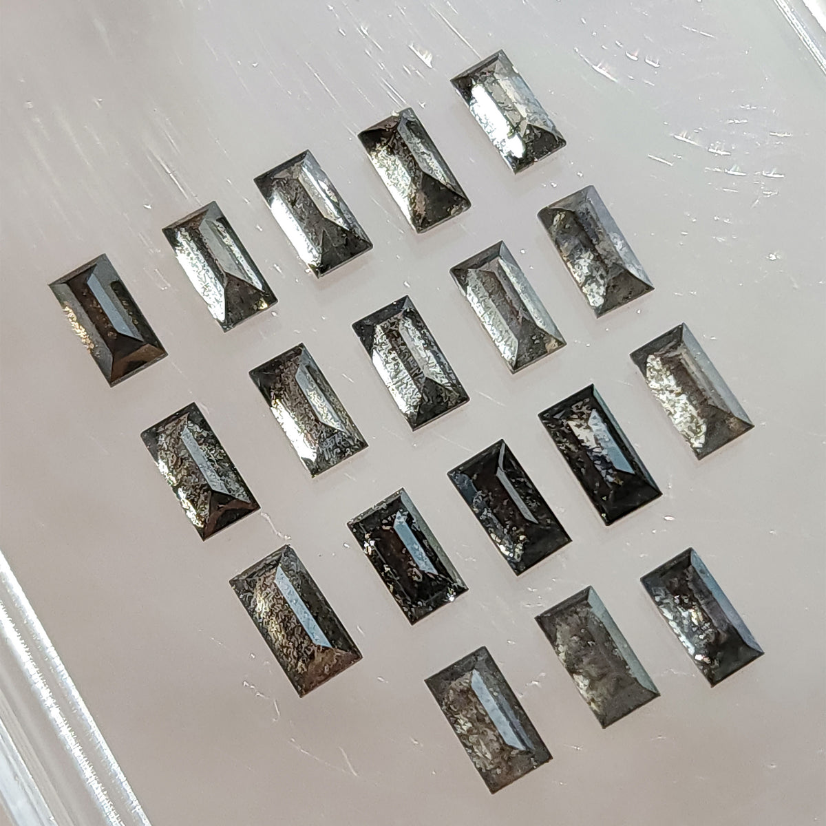 Angled view of salt and pepper diamond baguettes arranged diagonally on a light surface, showing gray and black inclusions and facet reflections.