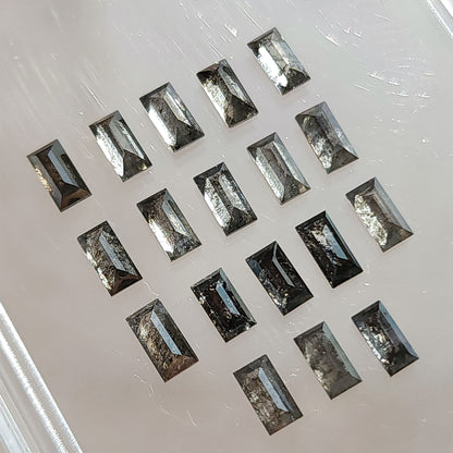 Angled view of salt and pepper diamond baguettes arranged diagonally on a light surface, showing gray and black inclusions and facet reflections.