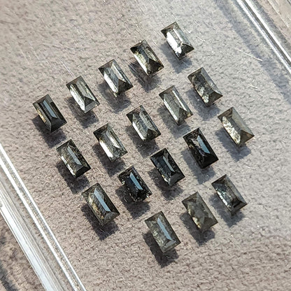 Salt and pepper diamond baguettes arranged in a clear tray on a textured surface, showing their straight sides and facet reflections.
