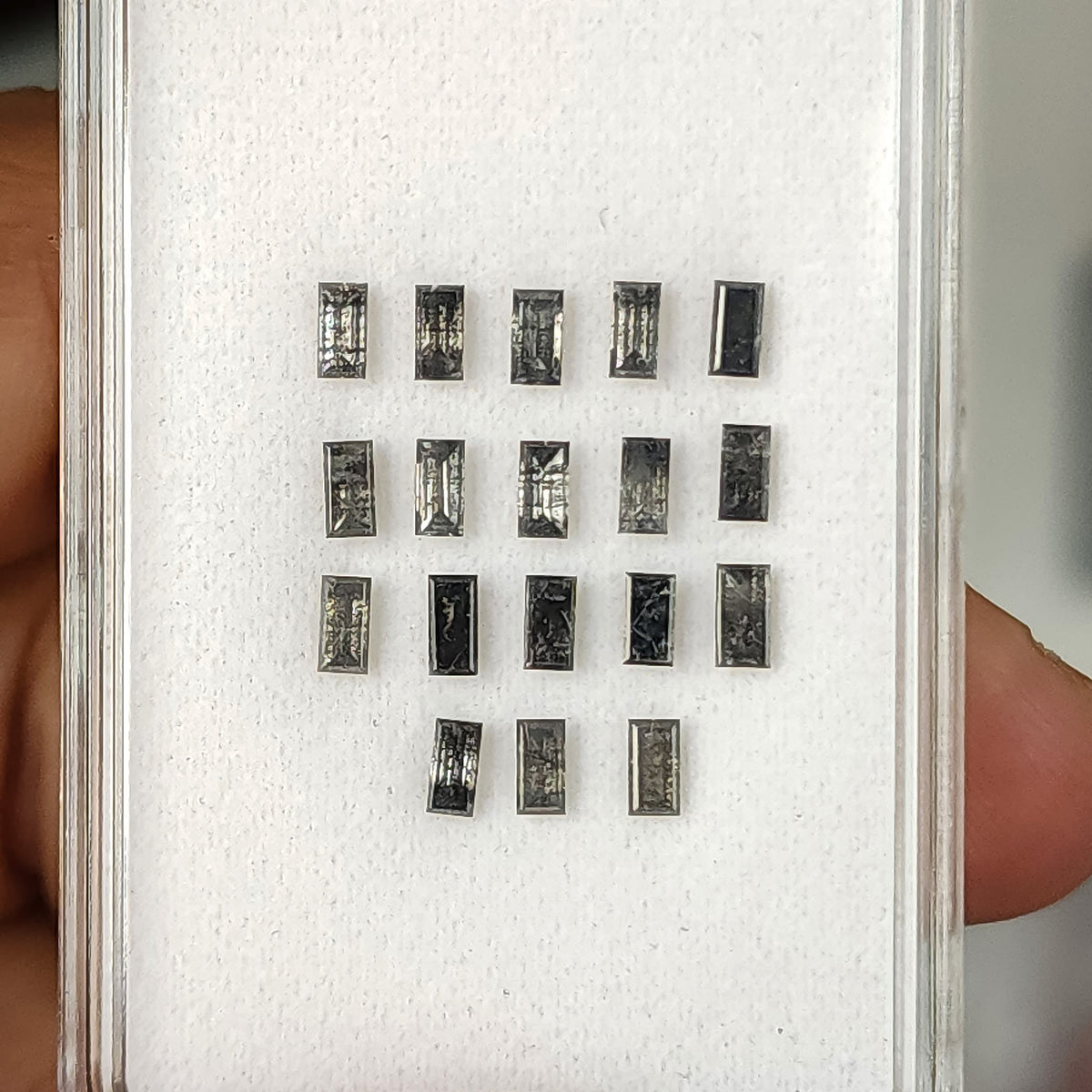 Rows of salt and pepper diamond baguettes on a white display pad held beside a finger for scale.