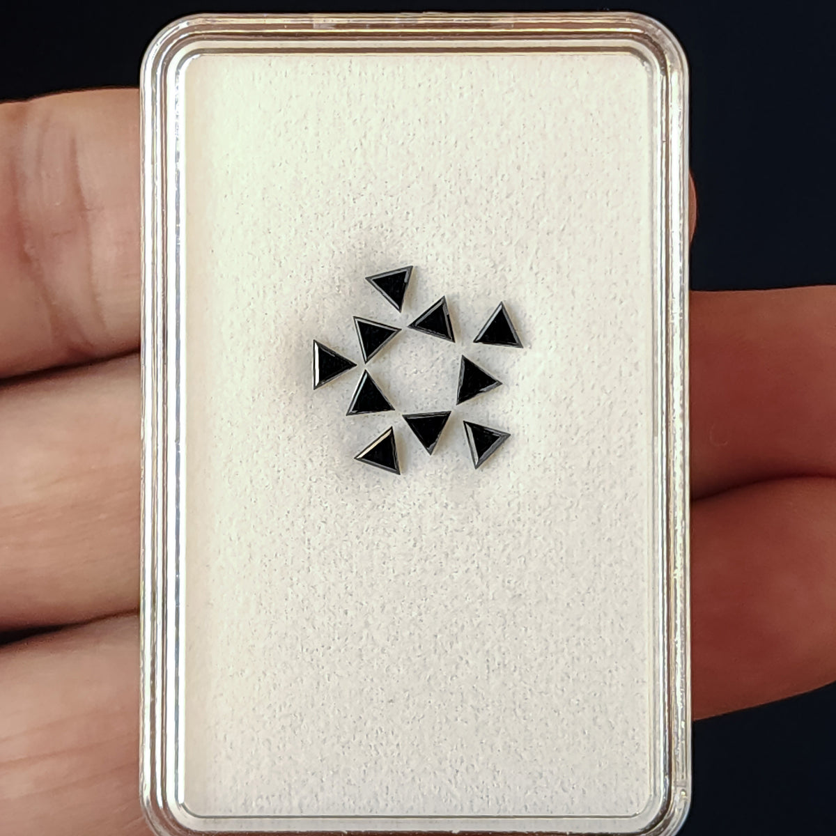 A small group of black triangular diamonds on a white cushion inside a gemstone box held by a hand.