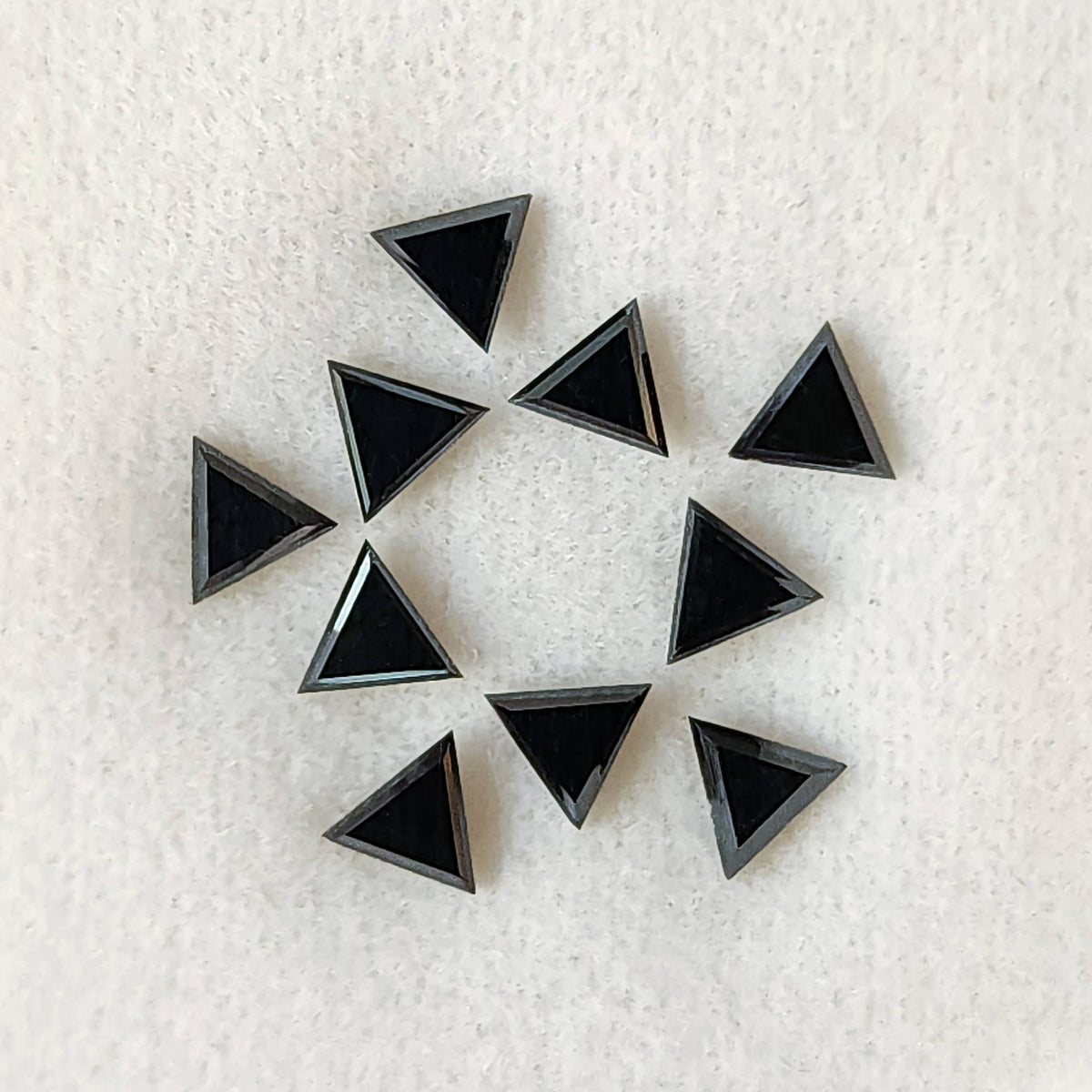 Ten black triangular diamonds on a white textured pad, showing broad flat triangular tables and narrow surrounding facets.