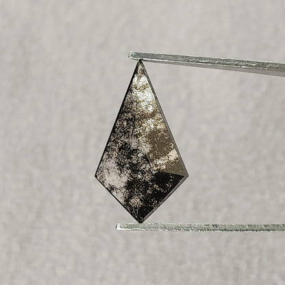 Face-up view of a speckled grey and black kite-shaped gemstone held in metal tweezers against a plain light grey background.