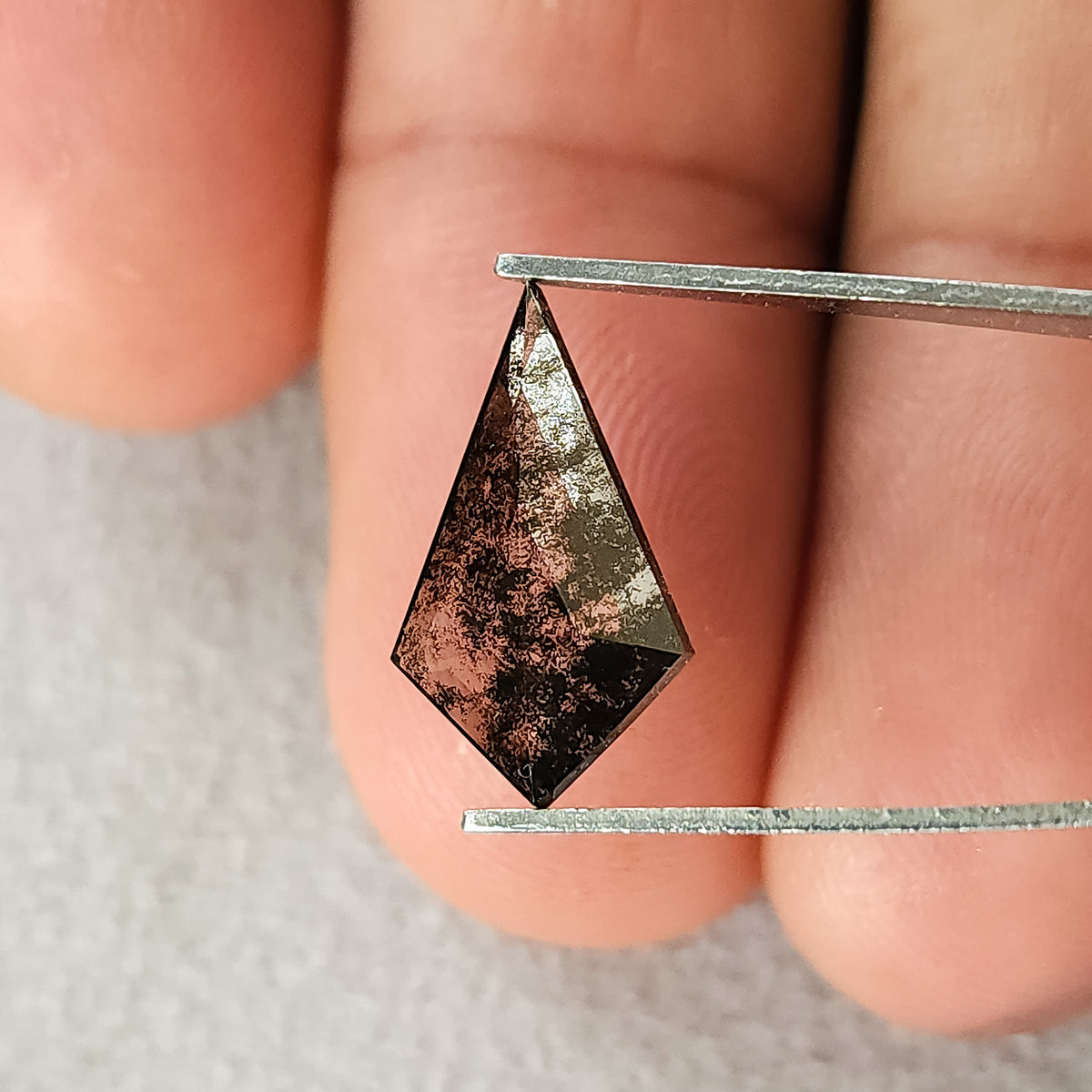Kite-shaped grey and black speckled gemstone held horizontally in tweezers, with out-of-focus fingers in the background.