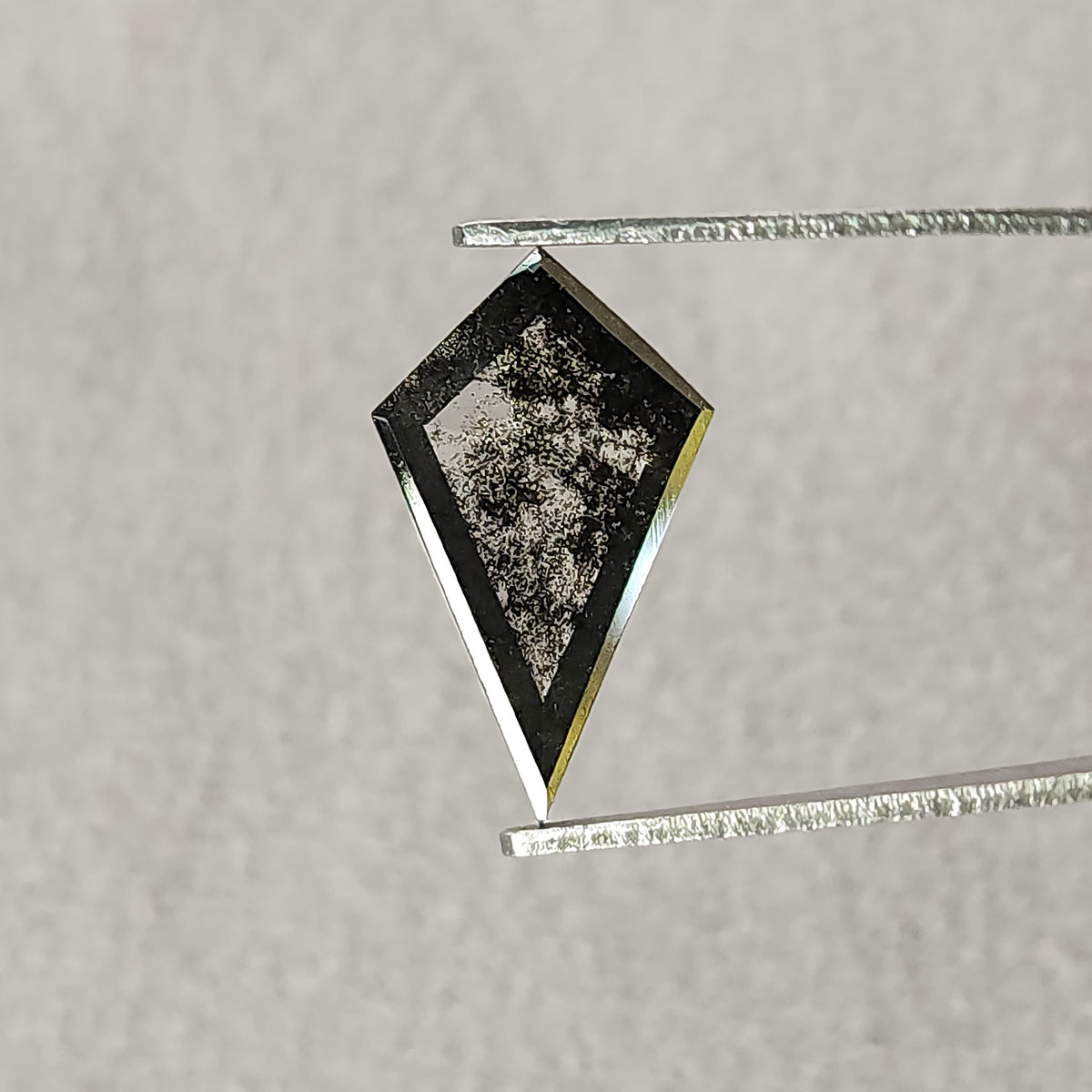 Tilted view of a dark grey and black kite-shaped gemstone in tweezers, showing a large flat reflective facet on a pale background.