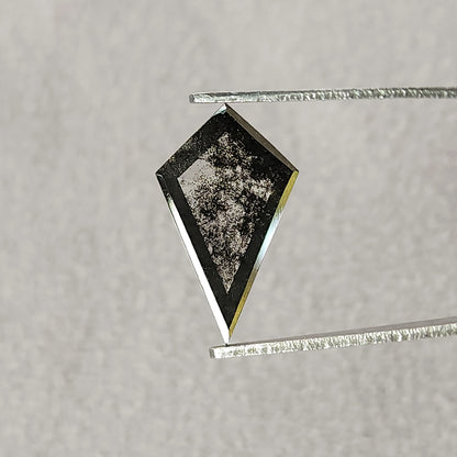 Tilted view of a dark grey and black kite-shaped gemstone in tweezers, showing a large flat reflective facet on a pale background.