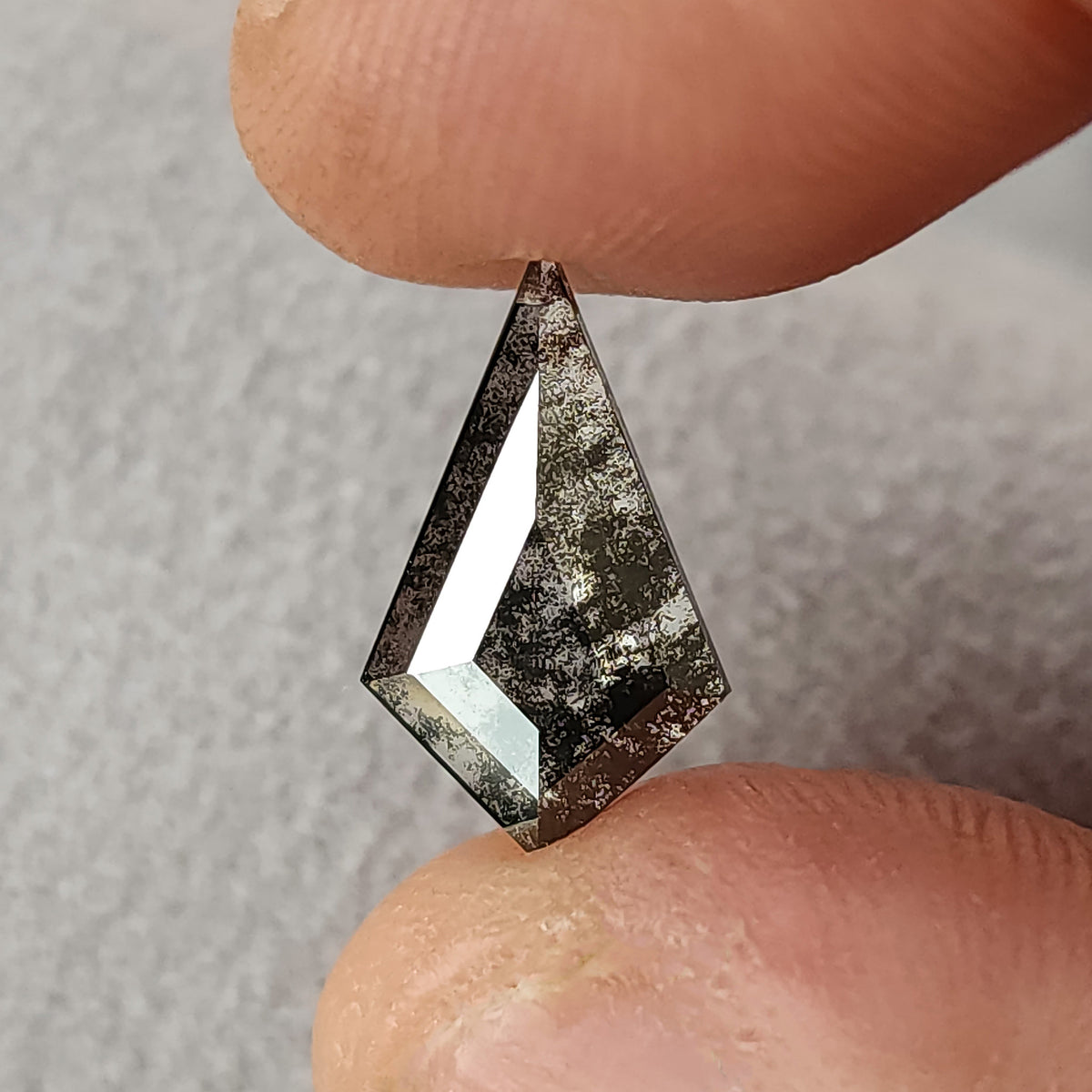 Grey and black speckled kite-shaped gemstone pinched between two fingers, highlighting sharp geometric facet reflections.