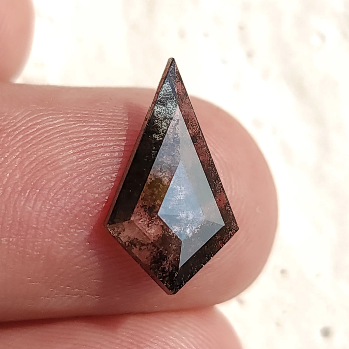 Close-up macro view of a speckled dark kite-shaped gemstone resting flat on a fingertip, showing clear geometric surface facets.