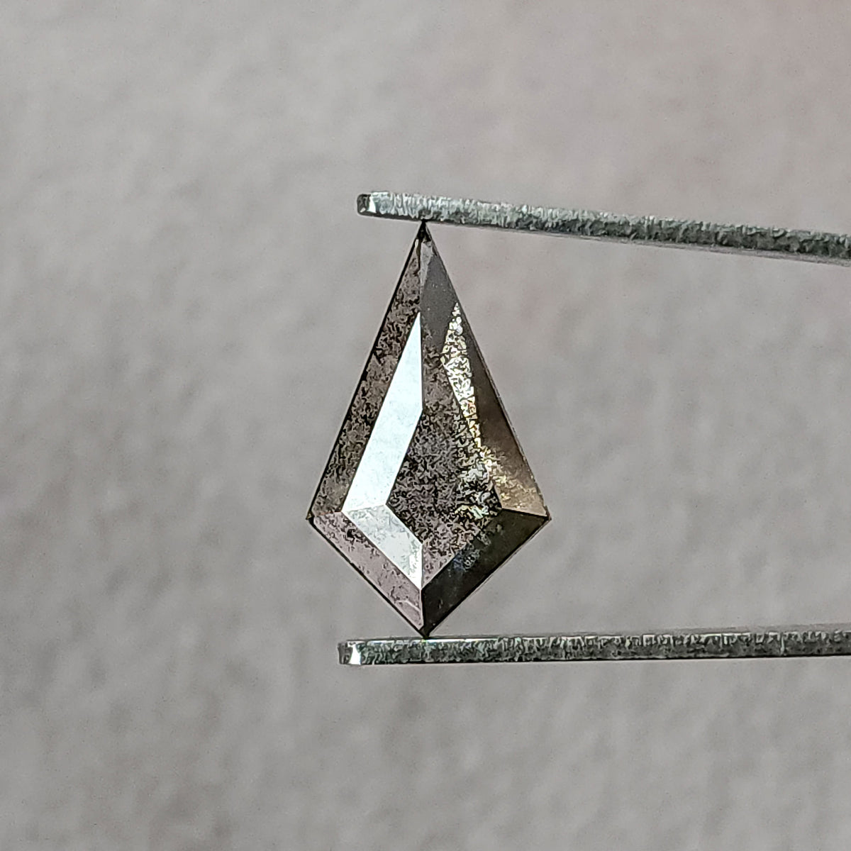 Face-up view of a kite-shaped salt and pepper lab-grown diamond held in metal tweezers against a light grey background.