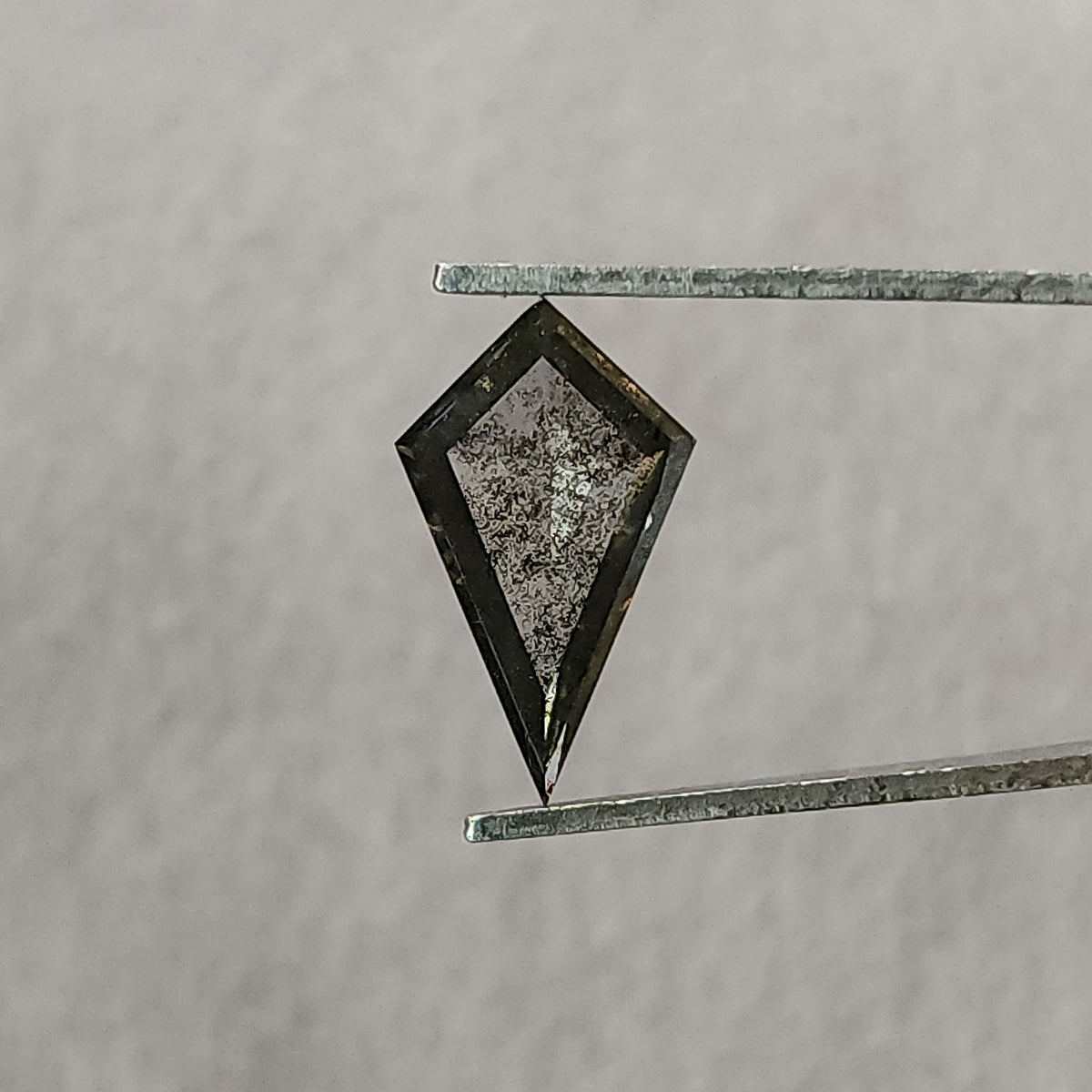 Face-up view of a kite-shaped salt and pepper lab-grown diamond held in tweezers against a plain light grey background.