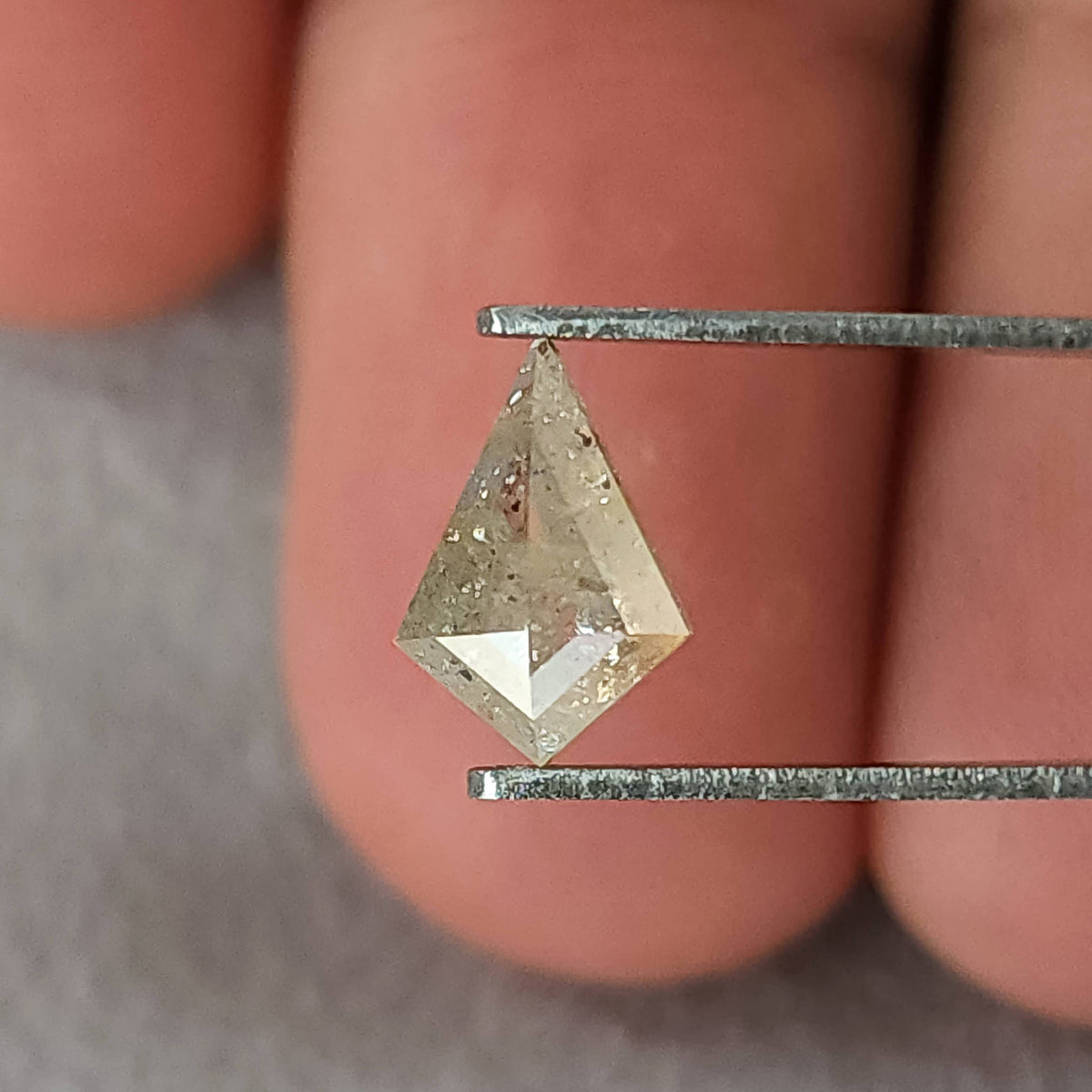 The kite-shaped natural diamond held in tweezers near fingers for scale, with pale champagne-gray reflections.