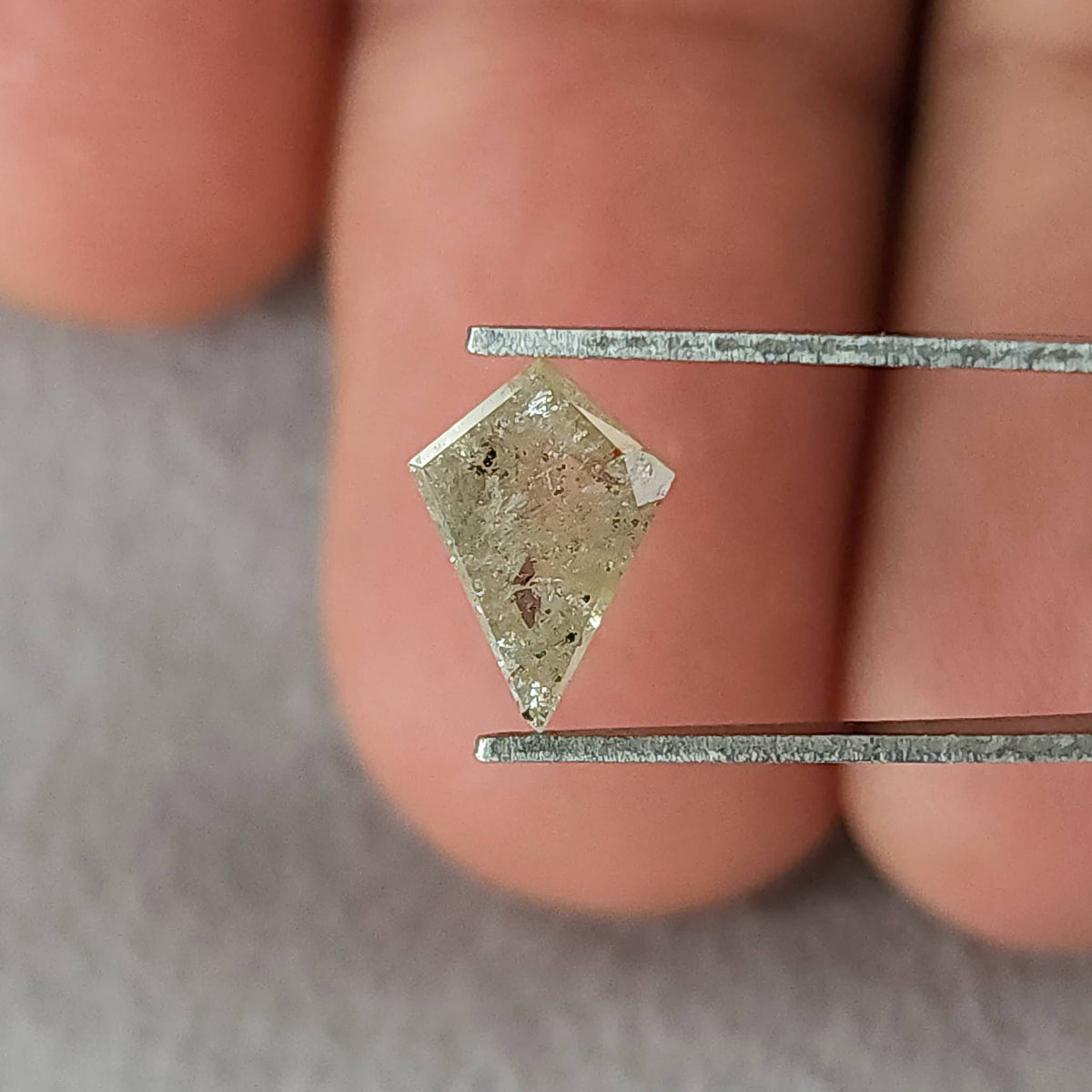 Opposite-side view of the kite-shaped natural diamond in tweezers, showing its inclusion pattern and warm gray reflections.
