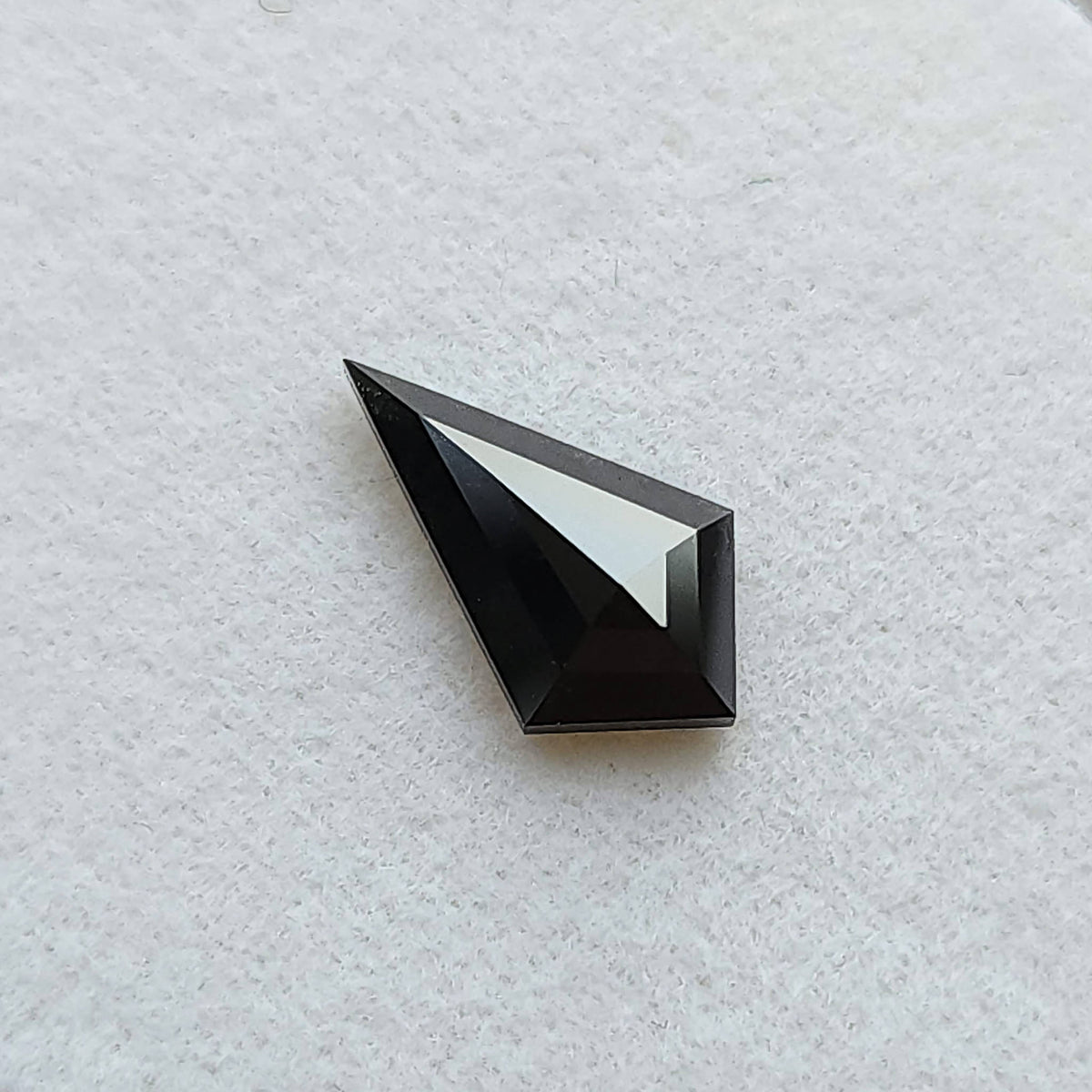 Vista inclinada de un diamante negro, talla cometa geométrica, sobre fondo blanco, resaltando un reflejo brillante en una faceta.