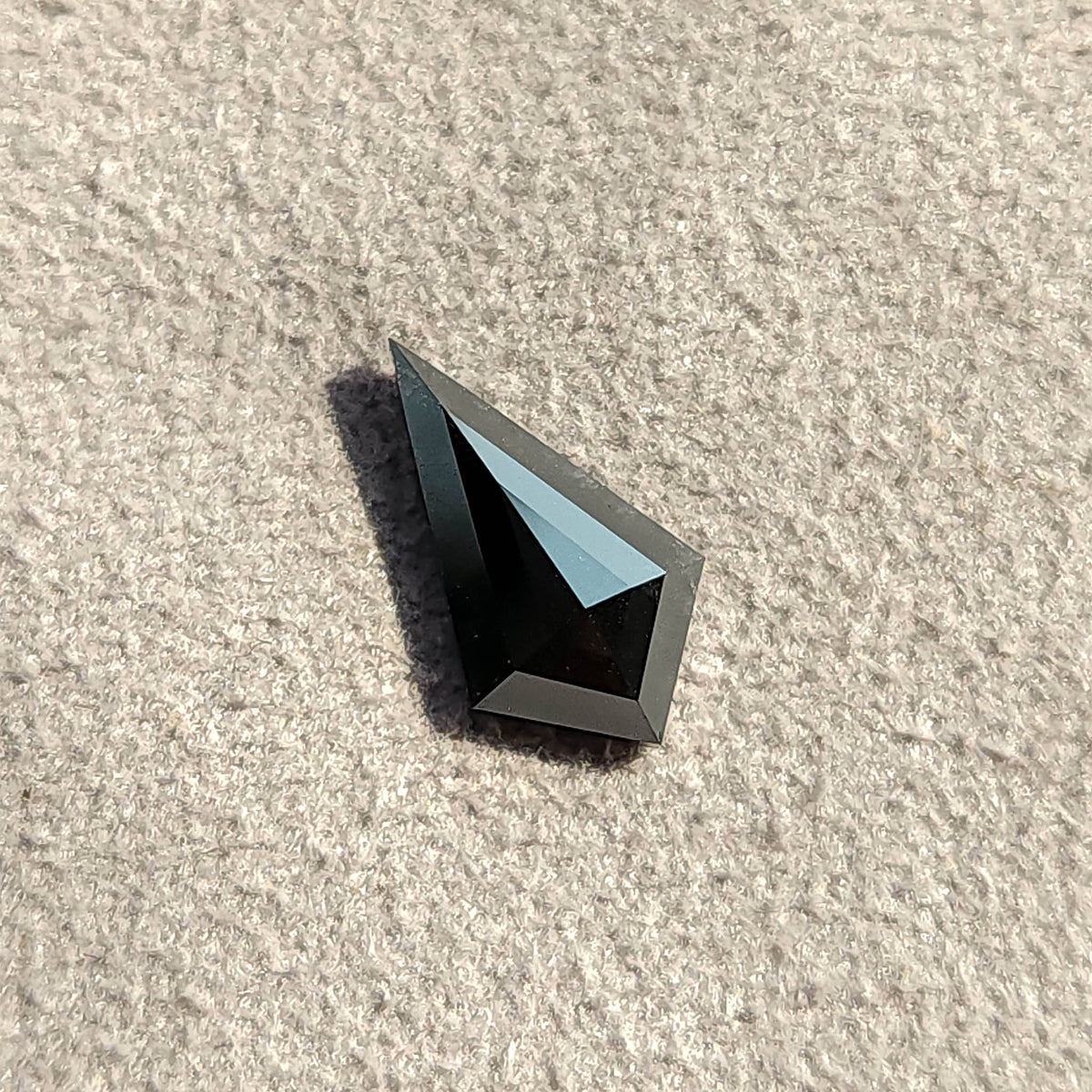 Vista inclinada de un diamante negro talla cometa bajo luz intensa, proyectando una sombra oscura sobre un fondo blanco.
