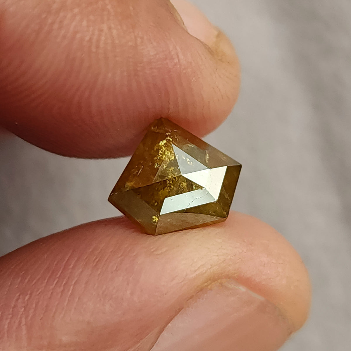 The shield-shaped diamond held near fingers for scale, showing its 9.4 × 8.2 mm face-up proportions.