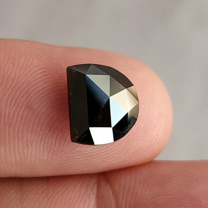 Dark brown D-shaped faceted gemstone resting flat on a fingertip, showing strong light and dark contrast across its surface.