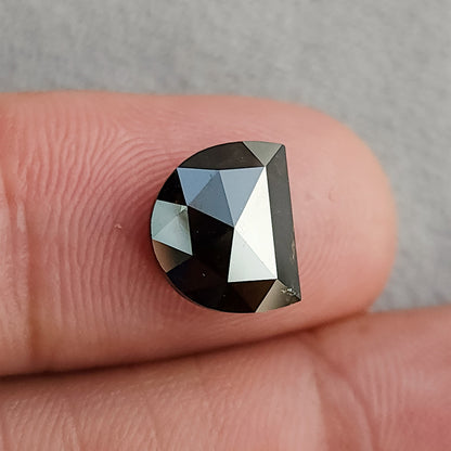 Face-up view resting on a finger, displaying a D-shaped, very dark brown gemstone with geometric facets against a pale background.