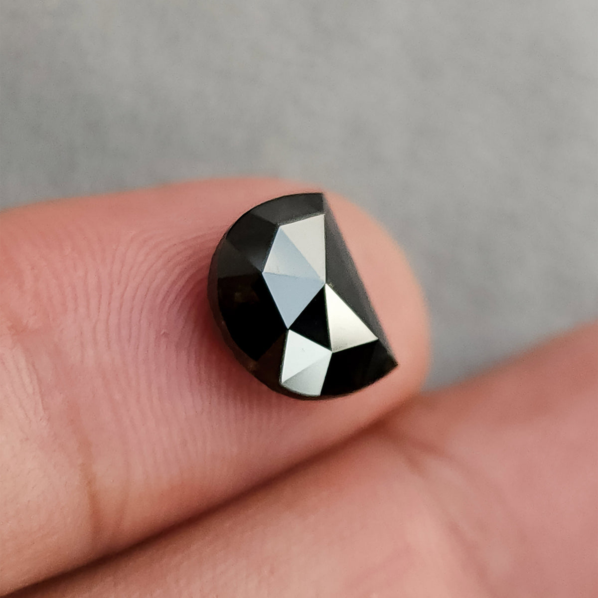 Angled face-up view of a dark, D-shaped gemstone sitting on a finger, catching bright light reflections on its triangular facets.