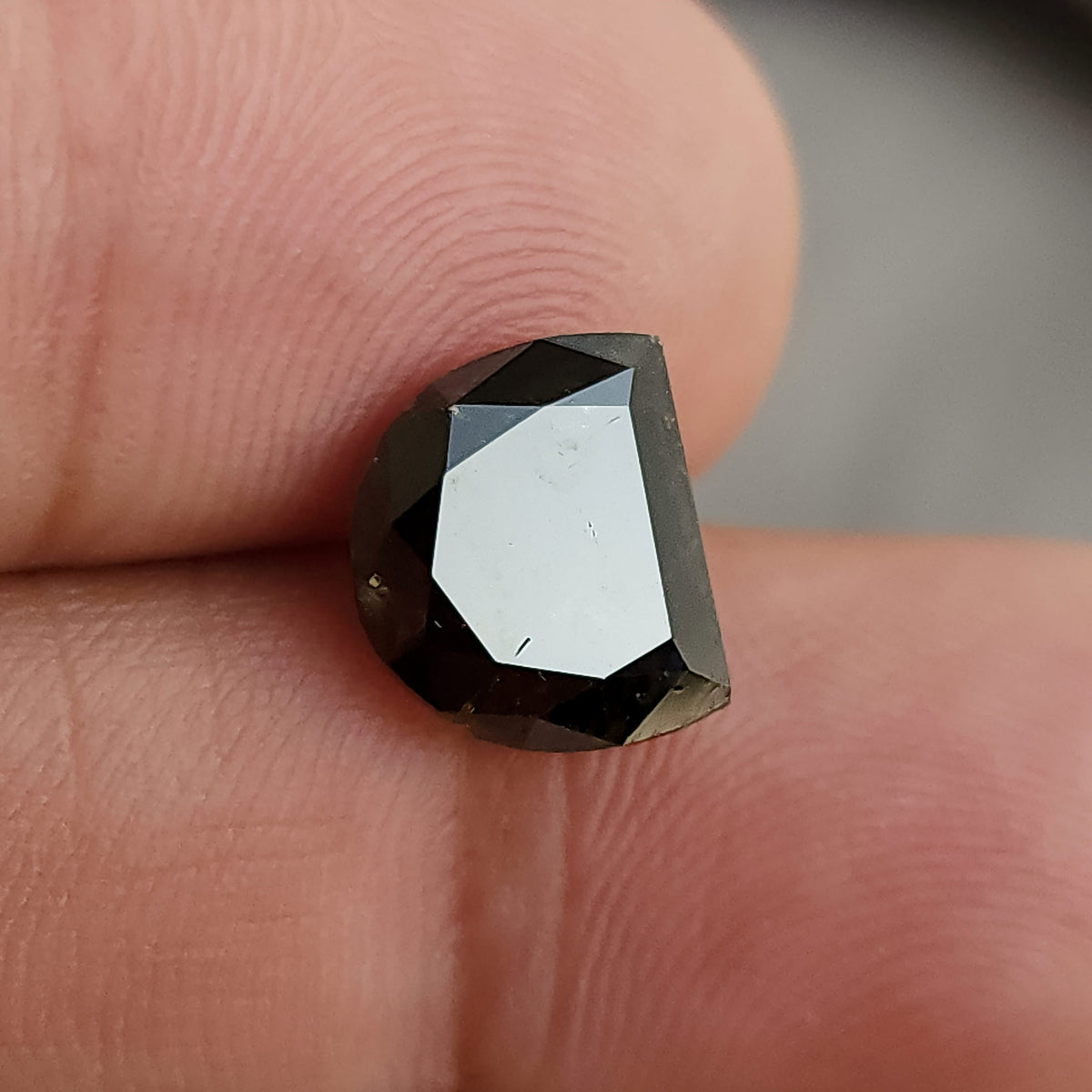 D-shaped dark gemstone lying flat on a finger, with a large, bright central facet reflection and dark surrounding step-like edges.
