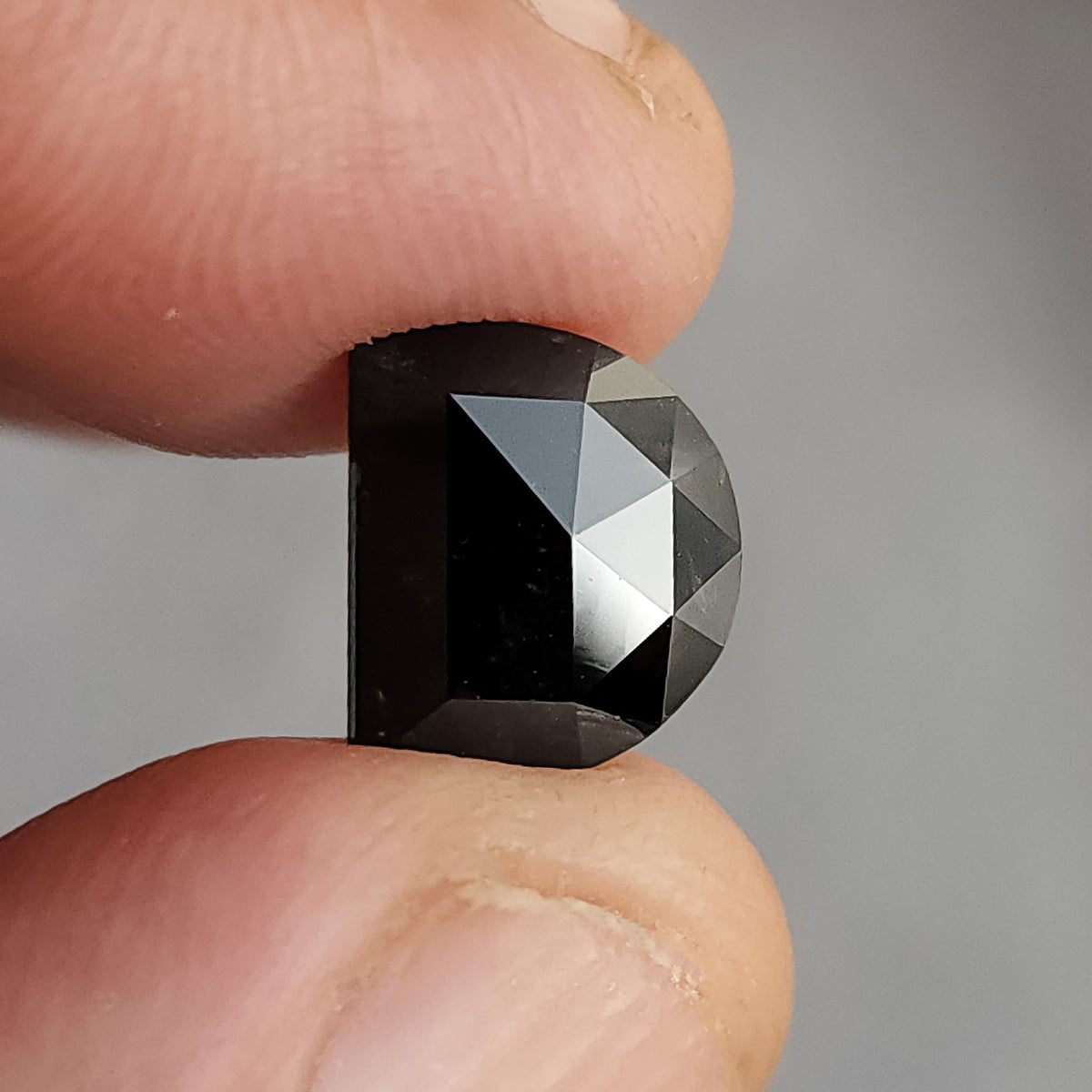 Profile view of the thick dark-brown D-shaped diamond held between two fingers, showing its thickness and side profile.